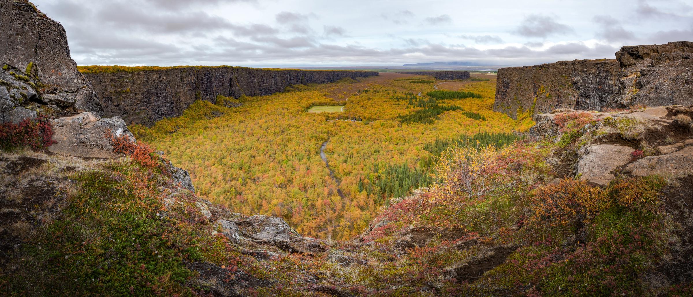 Panoramic view of an autumn valley filled with yellow and orange trees, flanked by dark cliffs, with a winding stream and small pond under a cloudy sky.