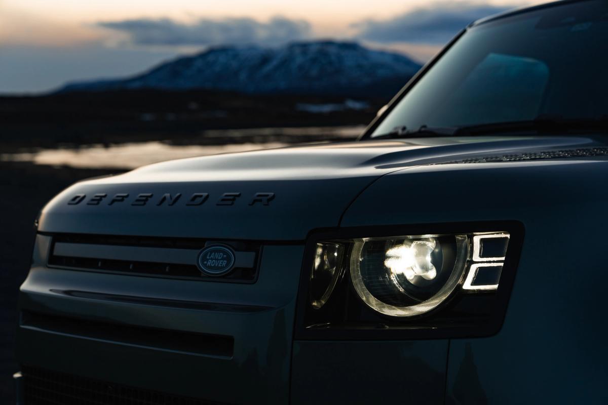Primer plano de la parrilla delantera y el faro de un Land Rover Defender al atardecer, con montañas al fondo, disponible en Go Car Rental Iceland