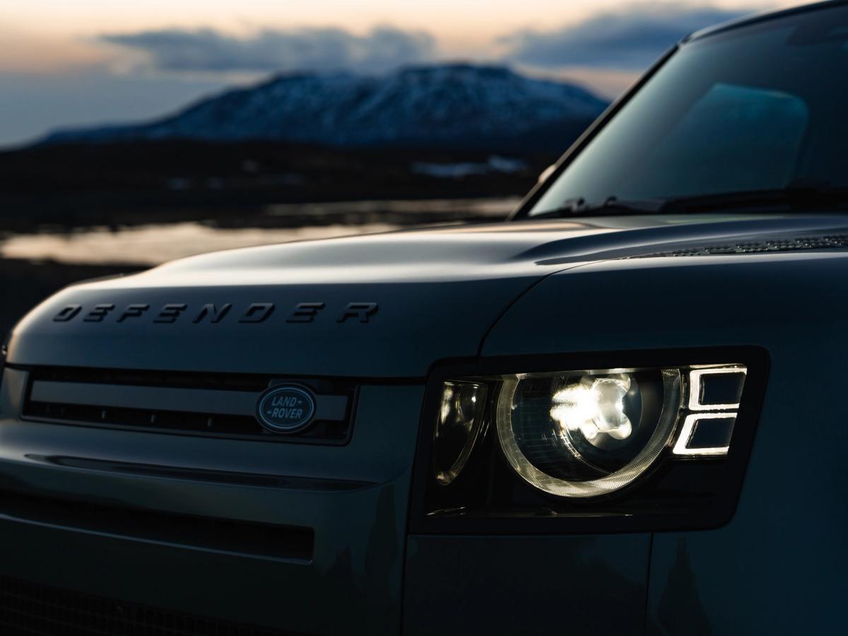 Primer plano del faro de un Land Rover Defender al anochecer Primer plano de la parrilla delantera y el faro de un Land Rover Defender al atardecer, con montañas al fondo, disponible en Go Car Rental Iceland