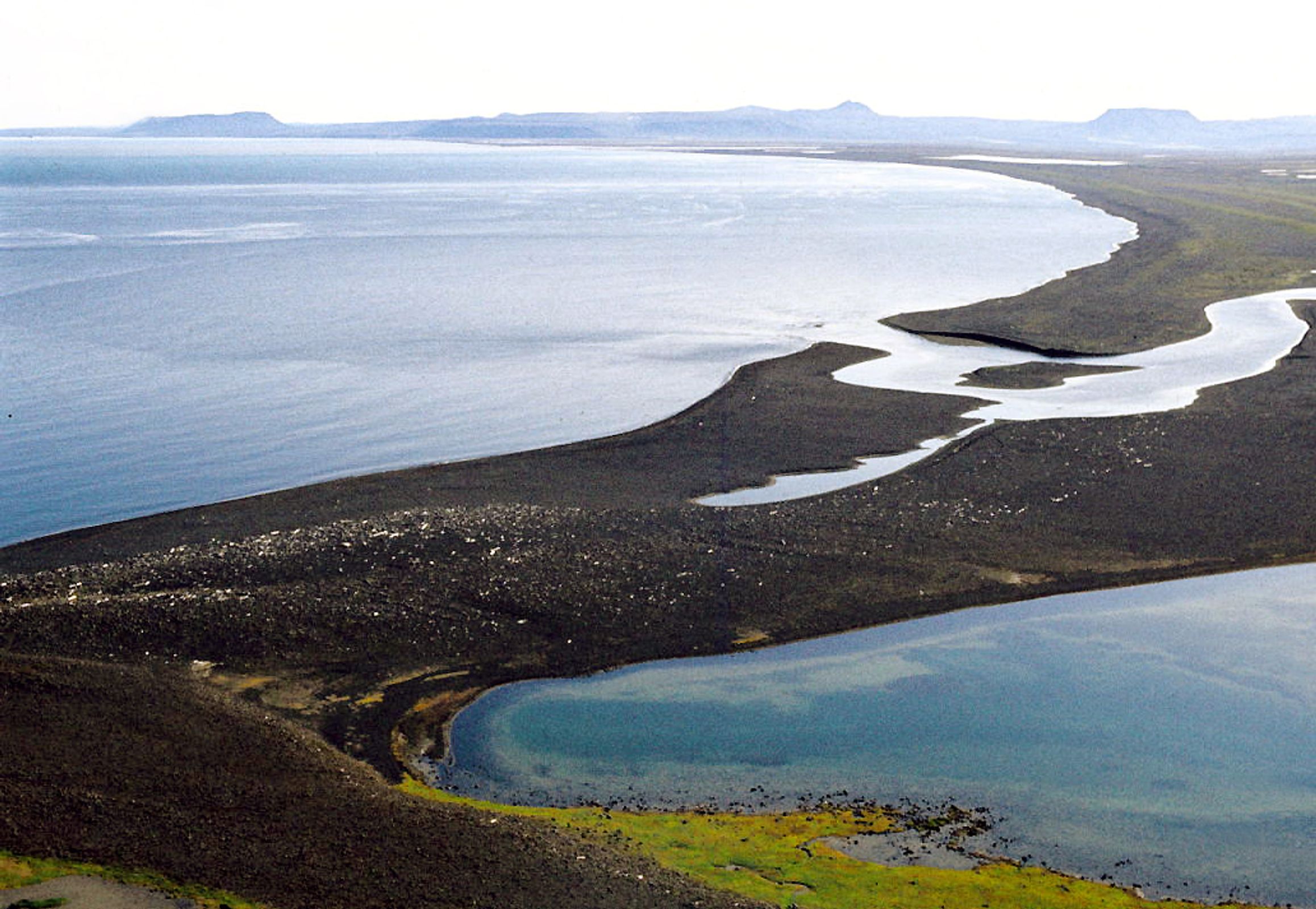 Aerial view of a dark, volcanic landform separating a light blue ocean from a winding river and a green-edged lagoon.