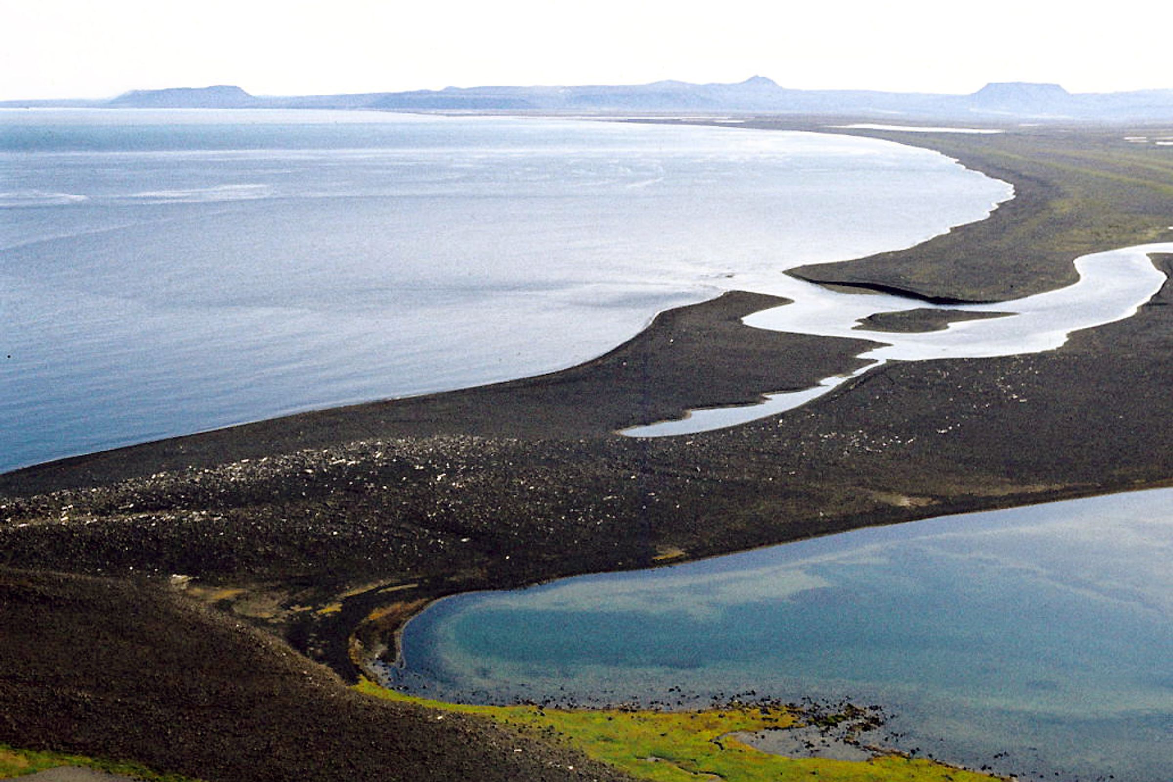 Aerial view of a dark, volcanic landform separating a light blue ocean from a winding river and a green-edged lagoon.