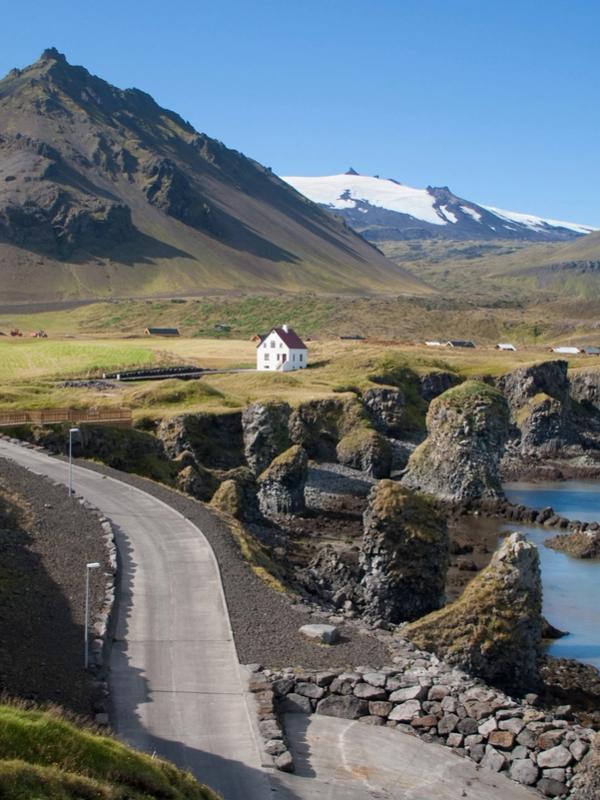 A winding road leads to a blue bay with sea stacks and a white house on a cliff, backed by a large dark mountain and a snow-capped glacier.