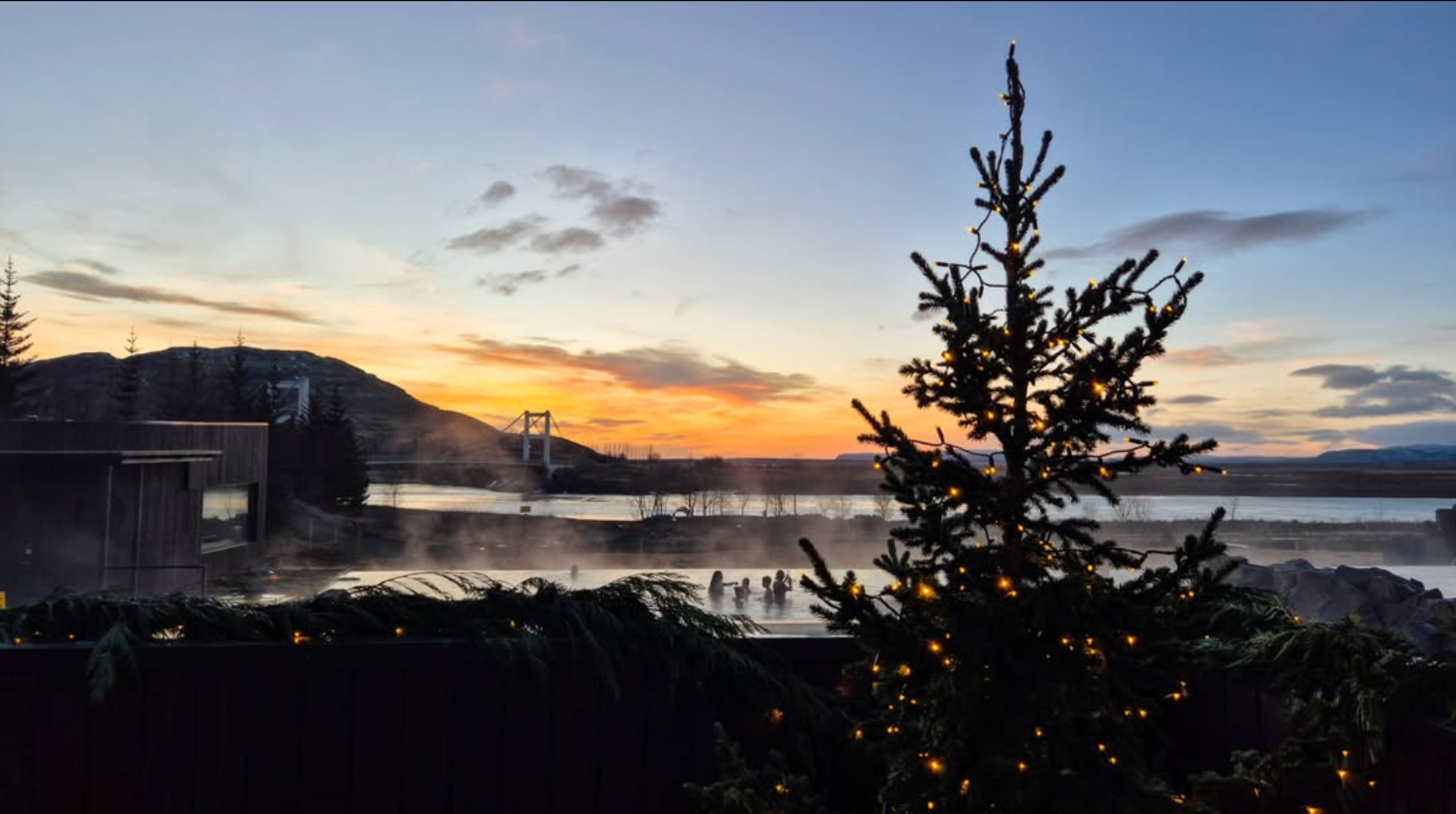 Un árbol de Navidad iluminado se alza frente a una fuente termal al aire libre humeante con gente dentro, todo enmarcado por un cielo de atardecer lleno de colores y un puente a lo lejos.