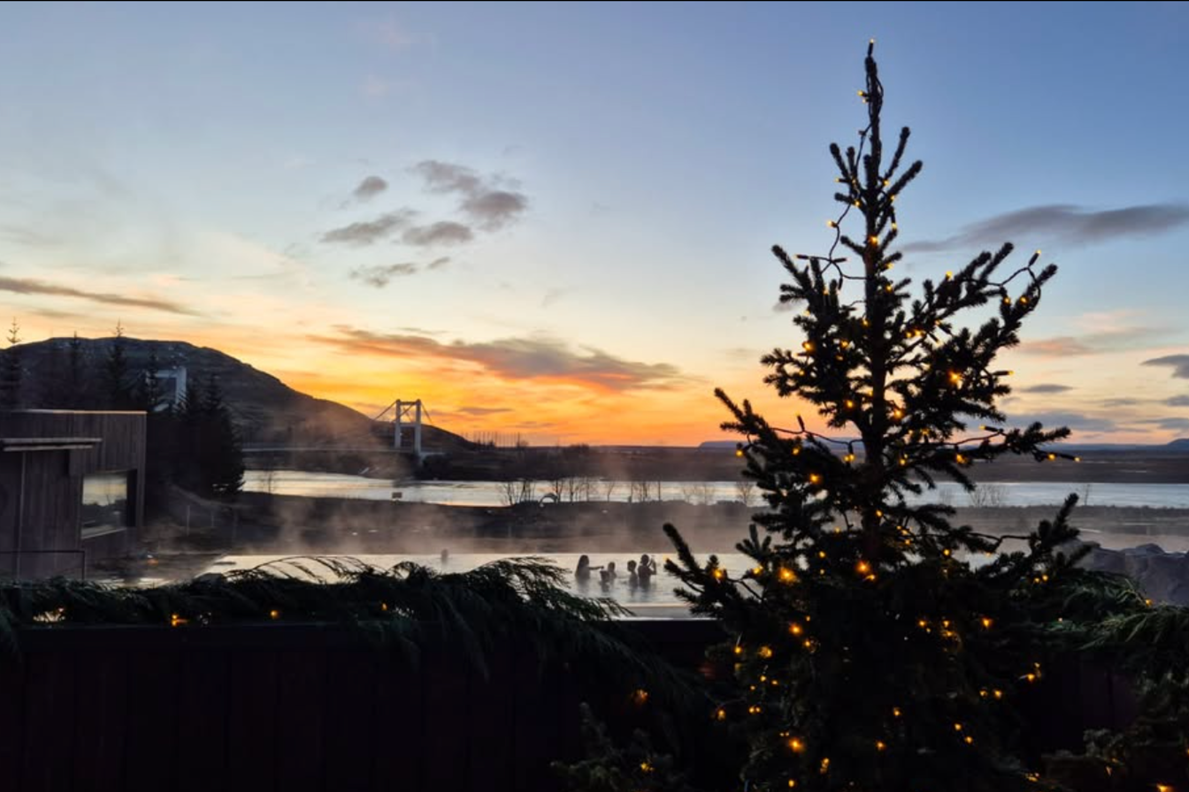 Un árbol de Navidad iluminado se alza frente a una fuente termal al aire libre humeante con gente dentro, todo enmarcado por un cielo de atardecer lleno de colores y un puente a lo lejos.