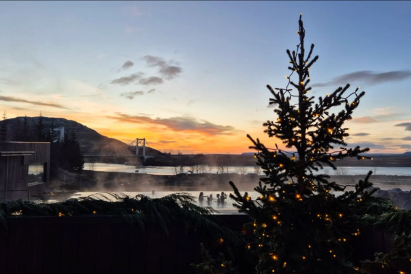 A Christmas tree with lights overlooks a steaming outdoor hot spring with people, set against a colorful sunset sky and distant bridge.