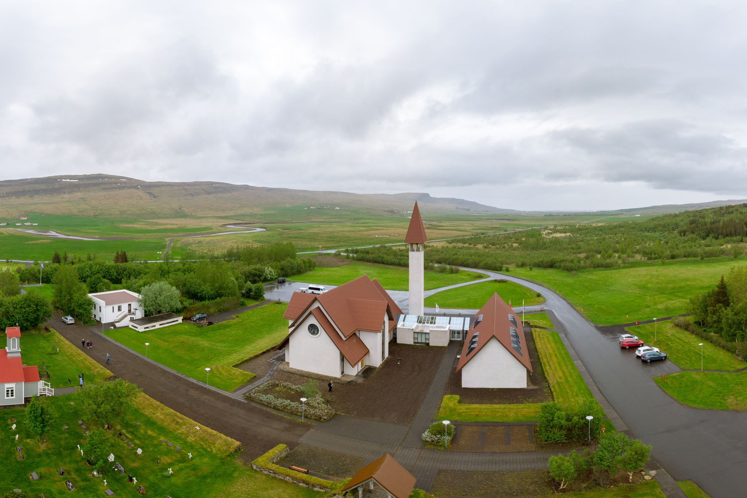 Aerial view of a village with a distinctive modern church and red-roofed buildings nestled in a vast green valley under an overcast sky.
