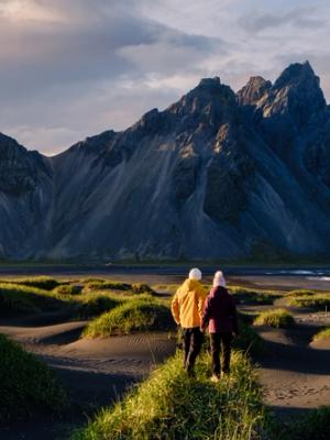 Two people on grassy black sand dunes face dramatic dark mountains and a black sand beach at sunset.