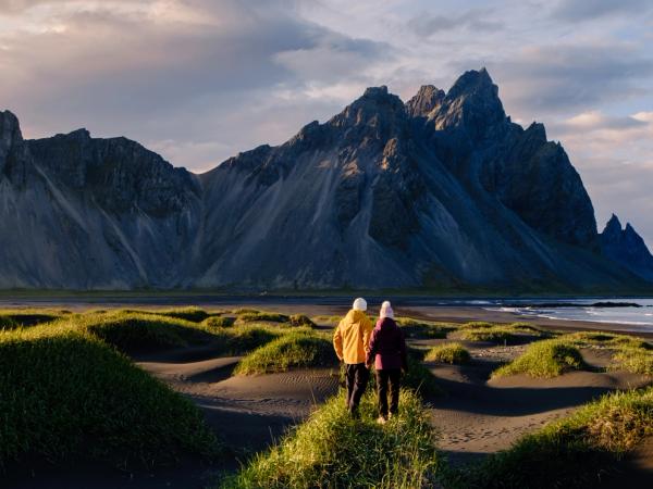 Two people on a grassy dune overlooking a black sand beach and jagged mountains under a golden sky.