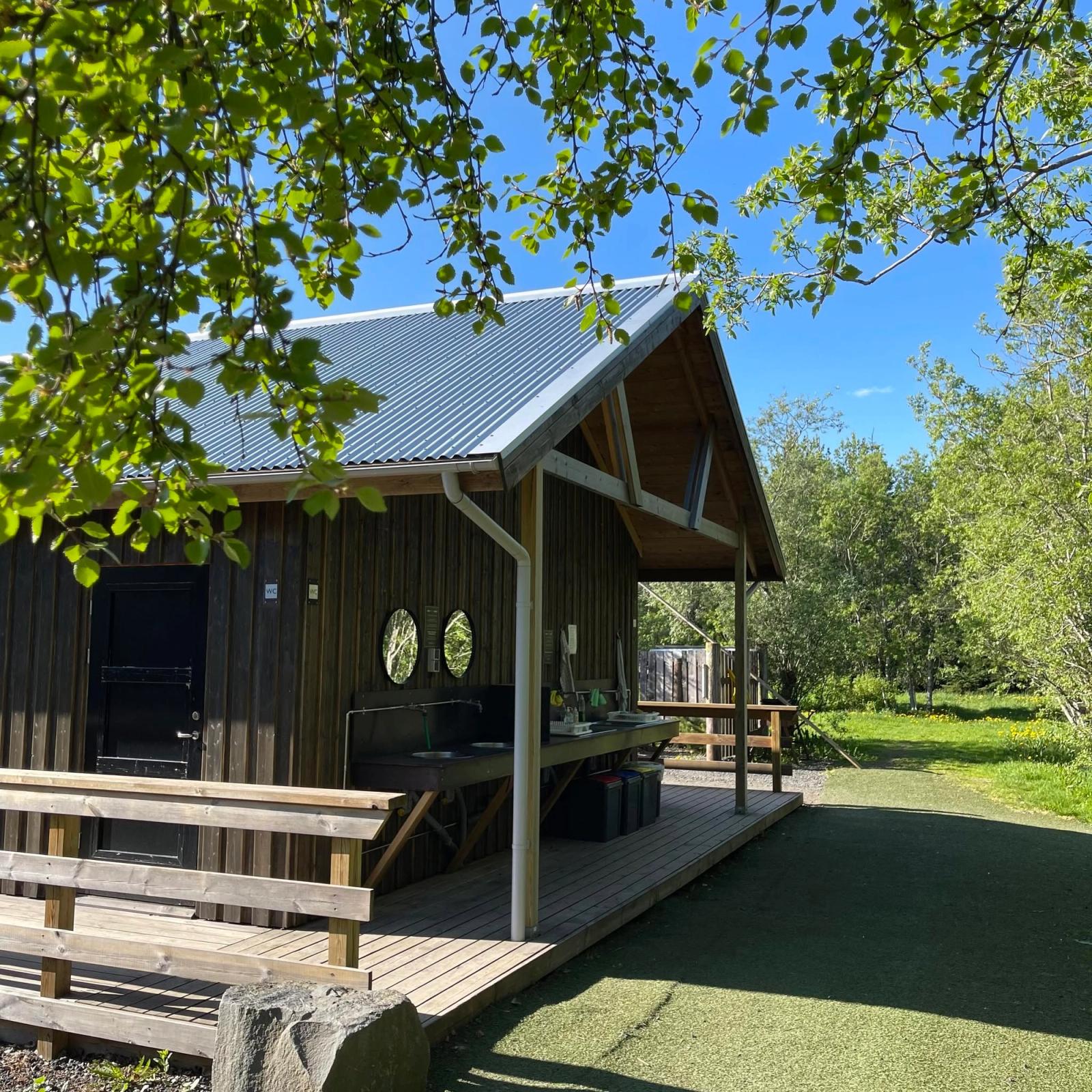 Wooden cabin with a metal roof, outdoor kitchen on a deck, surrounded by green trees under a blue sky.