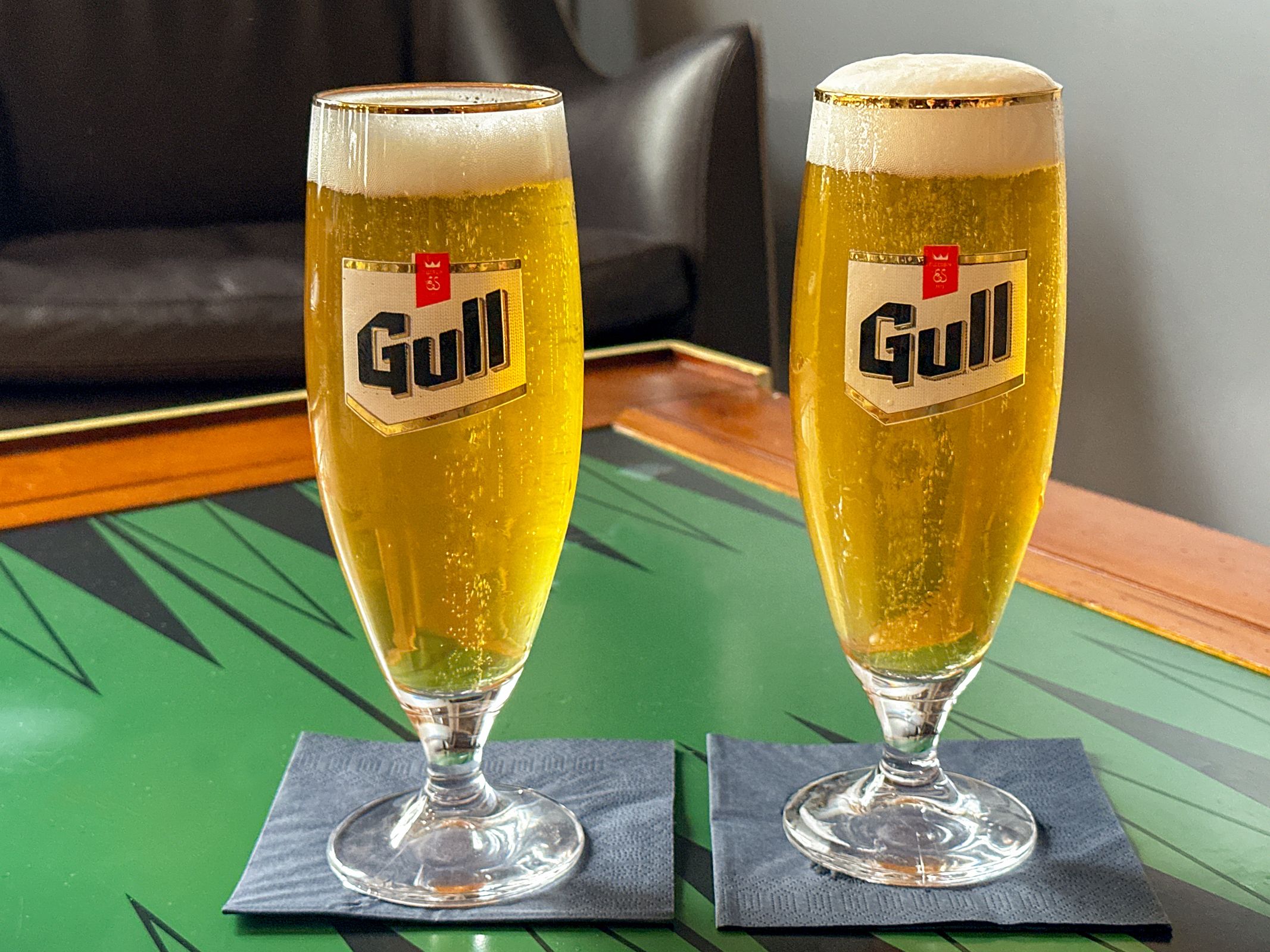 Two glasses of Gull beer with frothy heads on black napkins on a patterned table.