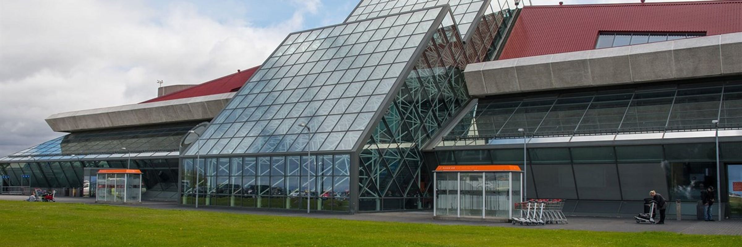 Modern airport terminal with a prominent glass facade, red roof, and green lawn.