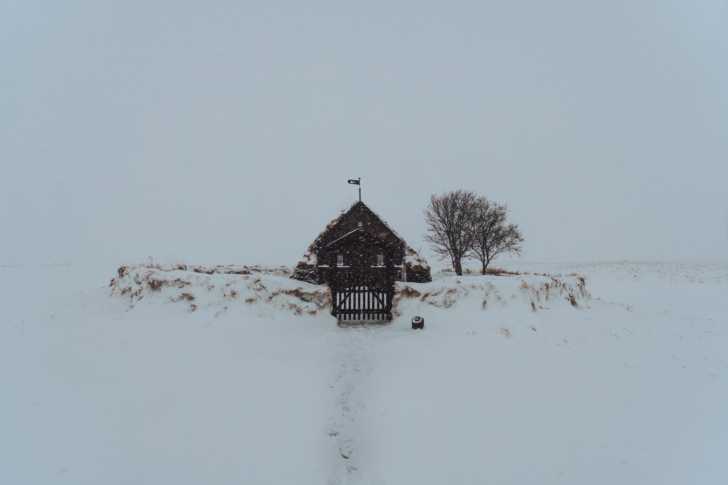 A small turf-roofed building with a gate and a bare tree stands in a vast, snowy landscape with falling snow and a path.