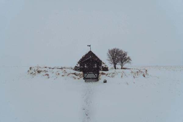 Un pequeño edificio con techo de césped, una puerta y un árbol desnudo se encuentra en un vasto paisaje nevado con nieve cayendo y un camino.