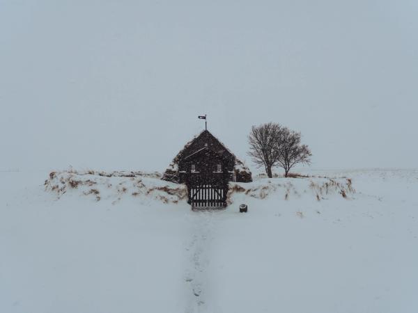 Un edificio oscuro con techo de césped, una puerta y un árbol desnudo en un campo desolado y cubierto de nieve con copos cayendo.