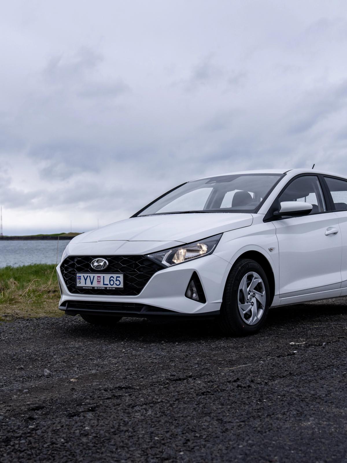 White Hyundai i20 on a gravel road by the sea under a cloudy sky.