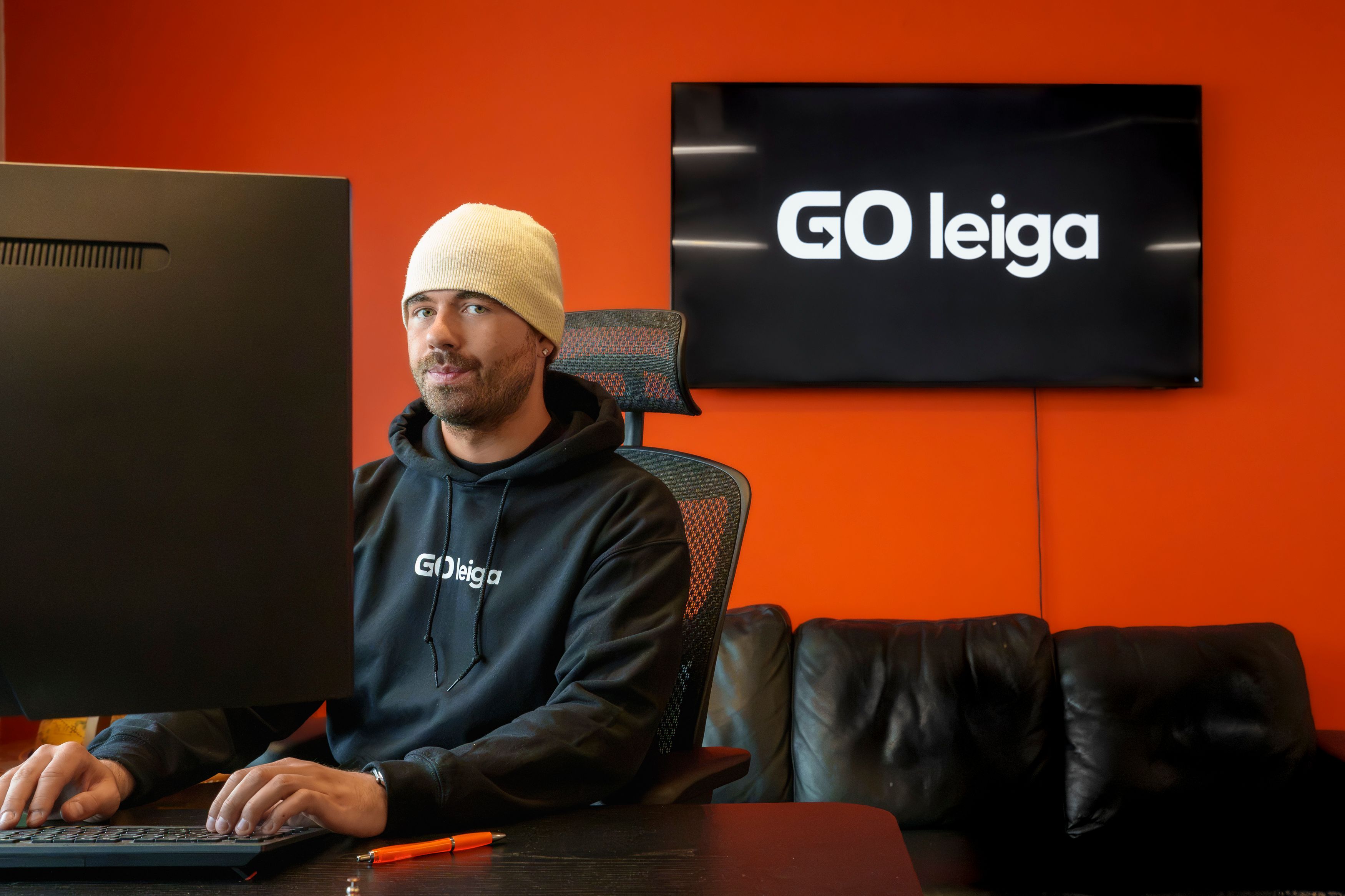 A man in a beanie and a "GO leiga" hoodie sits at a computer desk, with the "GO leiga" logo on a TV behind him.