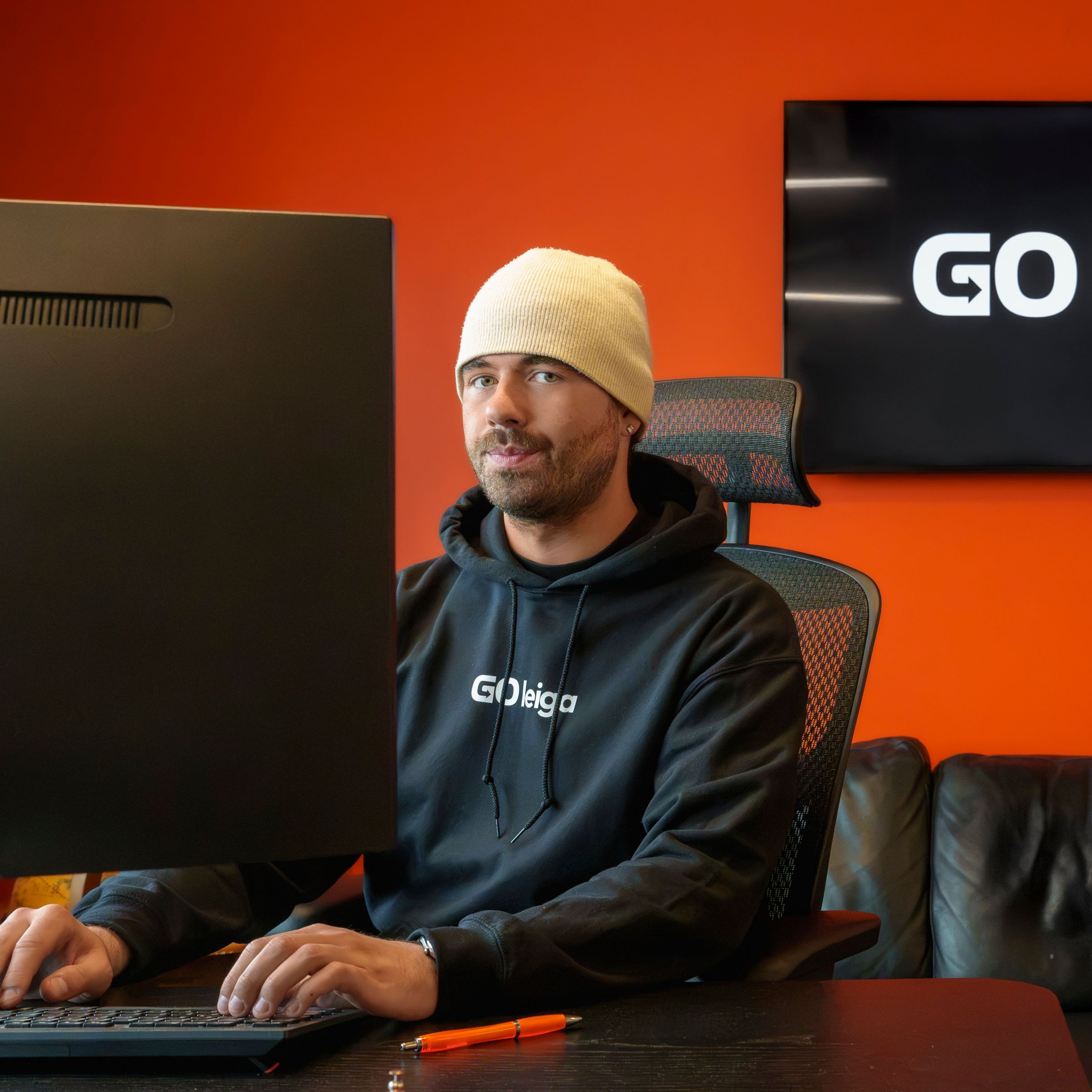 A man in a beanie and a "GO leiga" hoodie sits at a computer desk, with the "GO leiga" logo on a TV behind him.