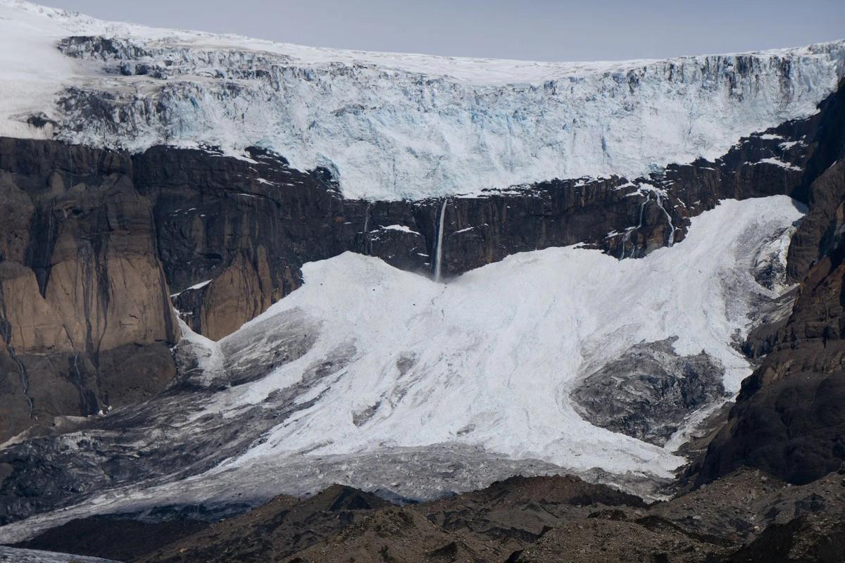 A glacier on a dark mountain cliff with multiple waterfalls cascading down to a debris-filled ice slope.