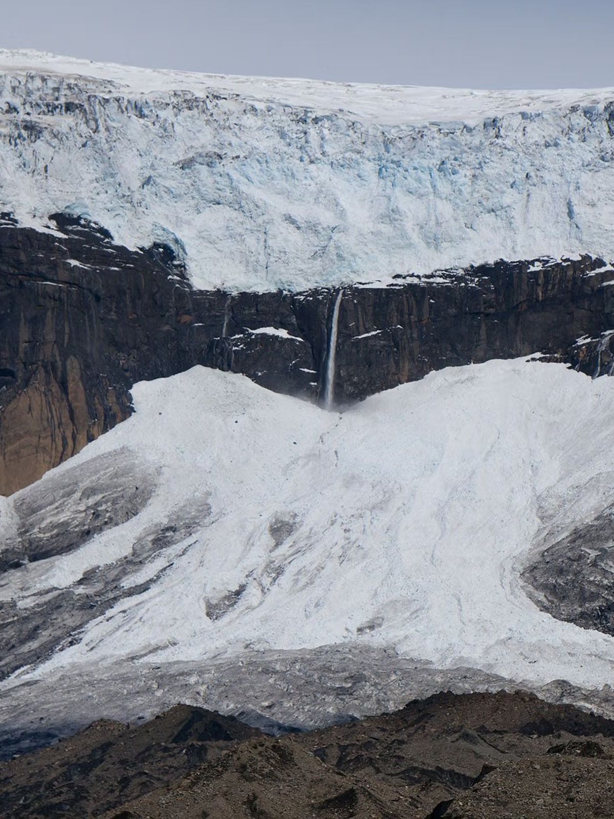 A glacier on a dark mountain cliff with multiple waterfalls cascading down to a debris-filled ice slope.