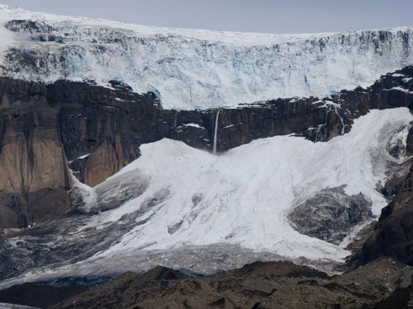 A glacier on a dark rock cliff with waterfalls cascading down into snowy, rocky terrain.