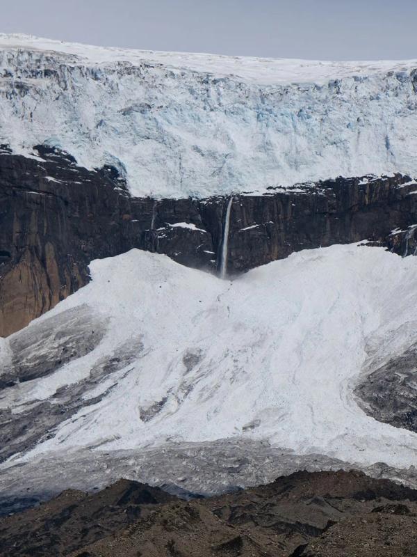 A glacier on a dark mountain cliff with multiple waterfalls cascading down to a debris-filled ice slope.