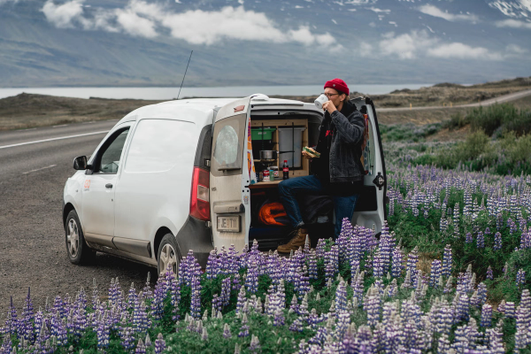 A man in a red beanie sits in the open back of a camper van, surrounded by purple lupines with a scenic mountain and lake backdrop.