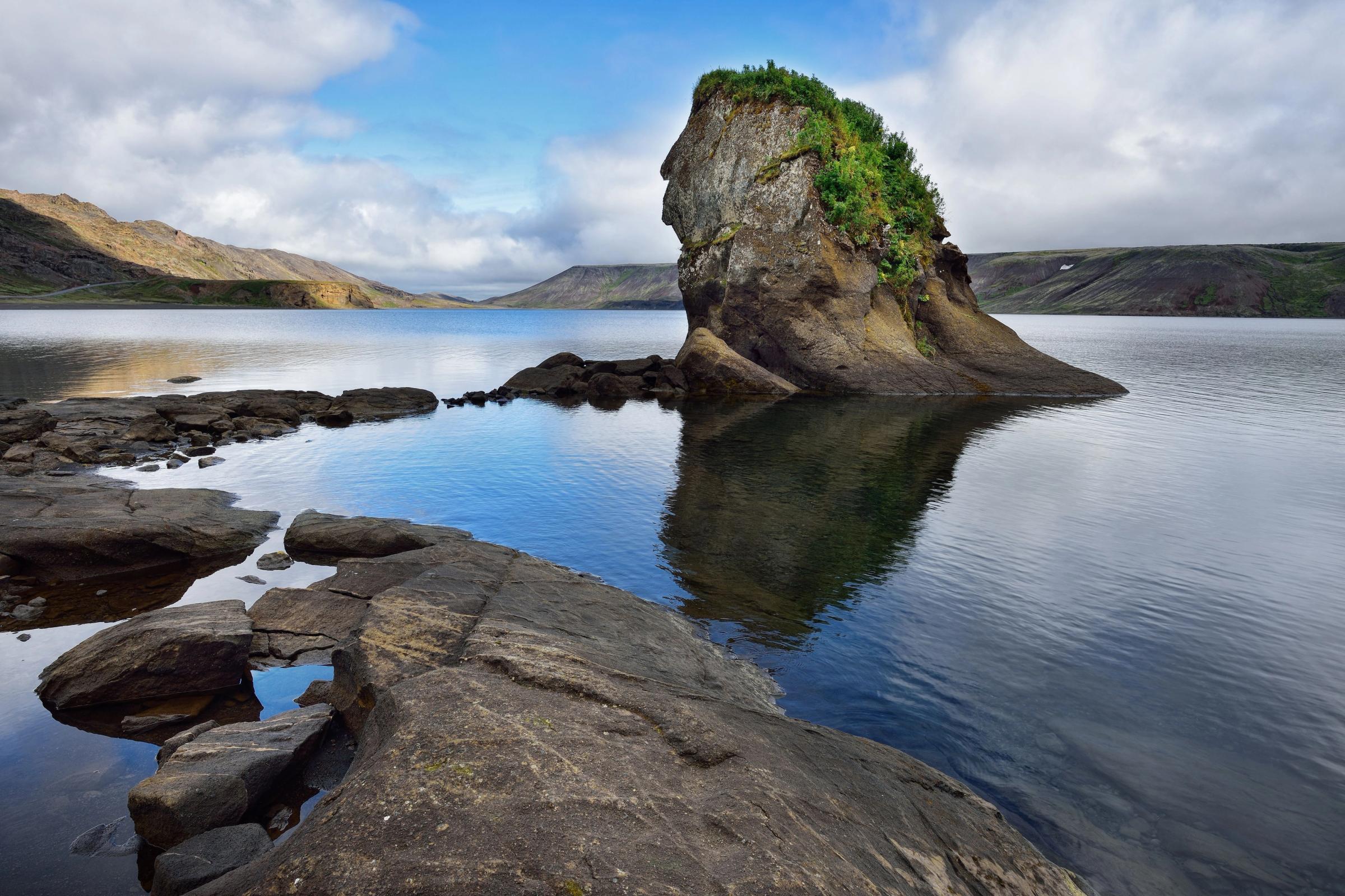 A rock formation resembling a human profile, topped with green vegetation, stands in a calm lake with rocky foreground and distant hills.