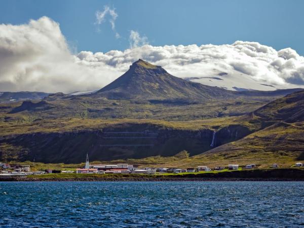 A coastal village nestled below green and rocky mountains, featuring a waterfall and a distant glacier under a partly cloudy sky.