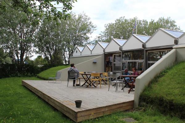 A modern building with a sawtooth roof and an outdoor wooden deck where people relax at tables, set into a green grassy slope with trees.