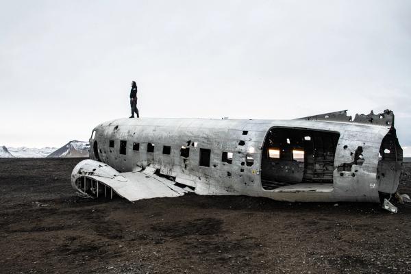 Person standing on the wreckage of a plane fuselage on a desolate black sand plain.