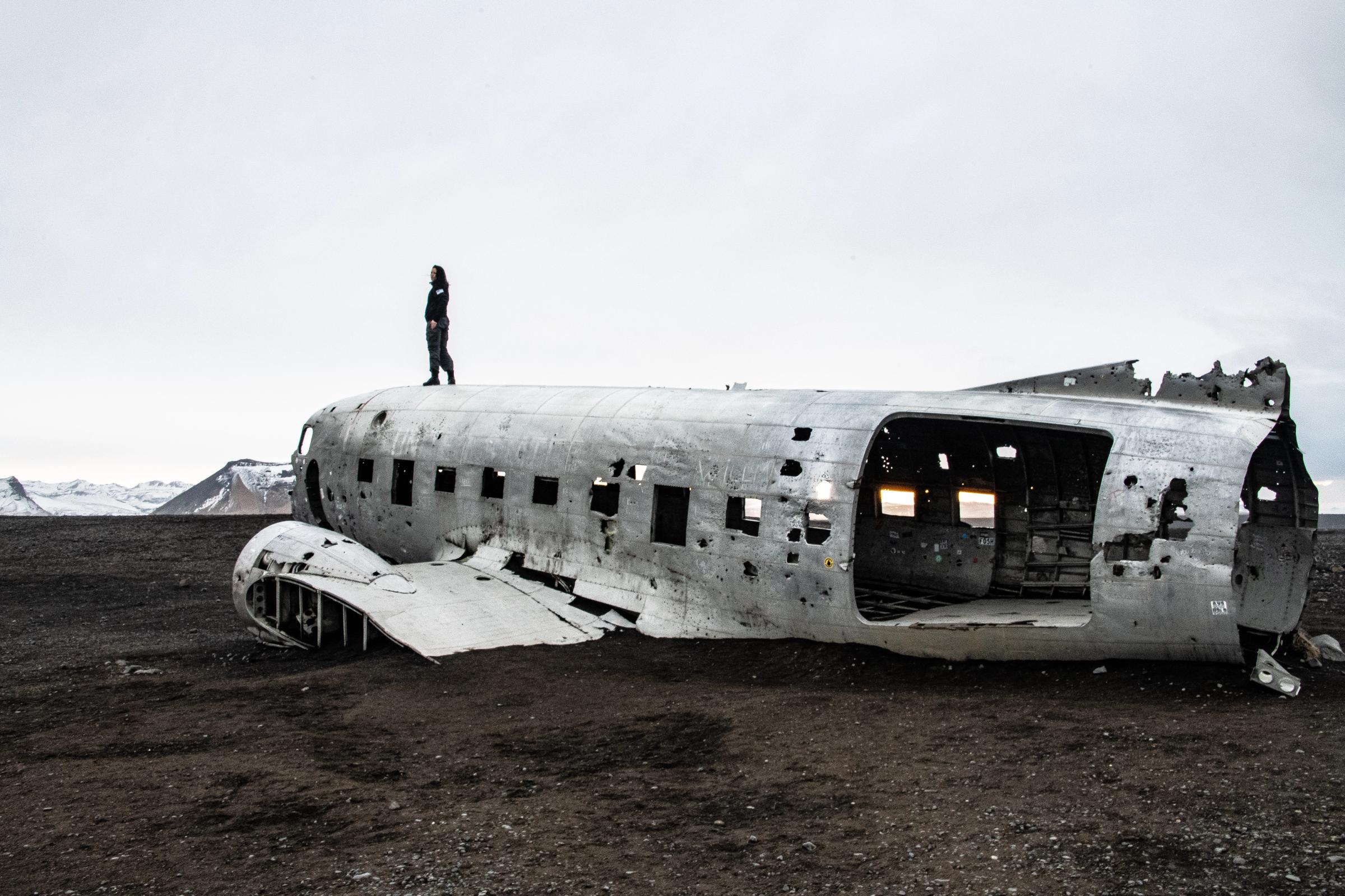 A person stands on the broken fuselage of a crashed airplane on a dark, desolate plain with distant mountains.