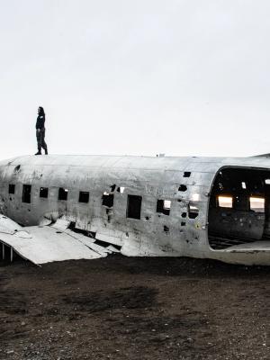 Person standing on the wreckage of a plane fuselage on a desolate black sand plain.