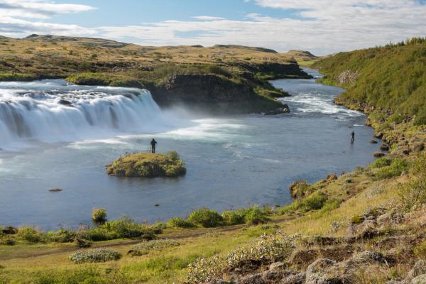 A scenic waterfall cascades into a river where two people fish, surrounded by green and brown hills under a blue sky.
