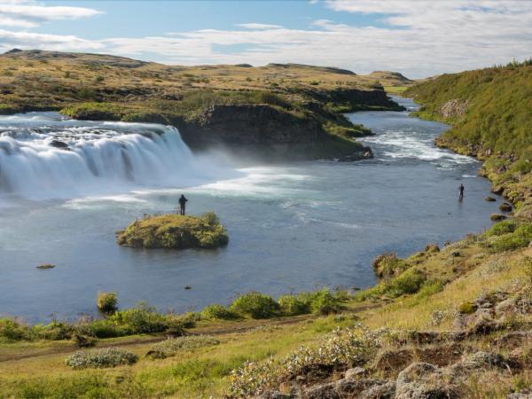 A river scene with a powerful waterfall, two people fishing, and rolling hills.