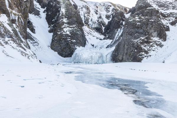 A snowy valley with a frozen stream leading to a large ice formation at the base of snow-covered cliffs.