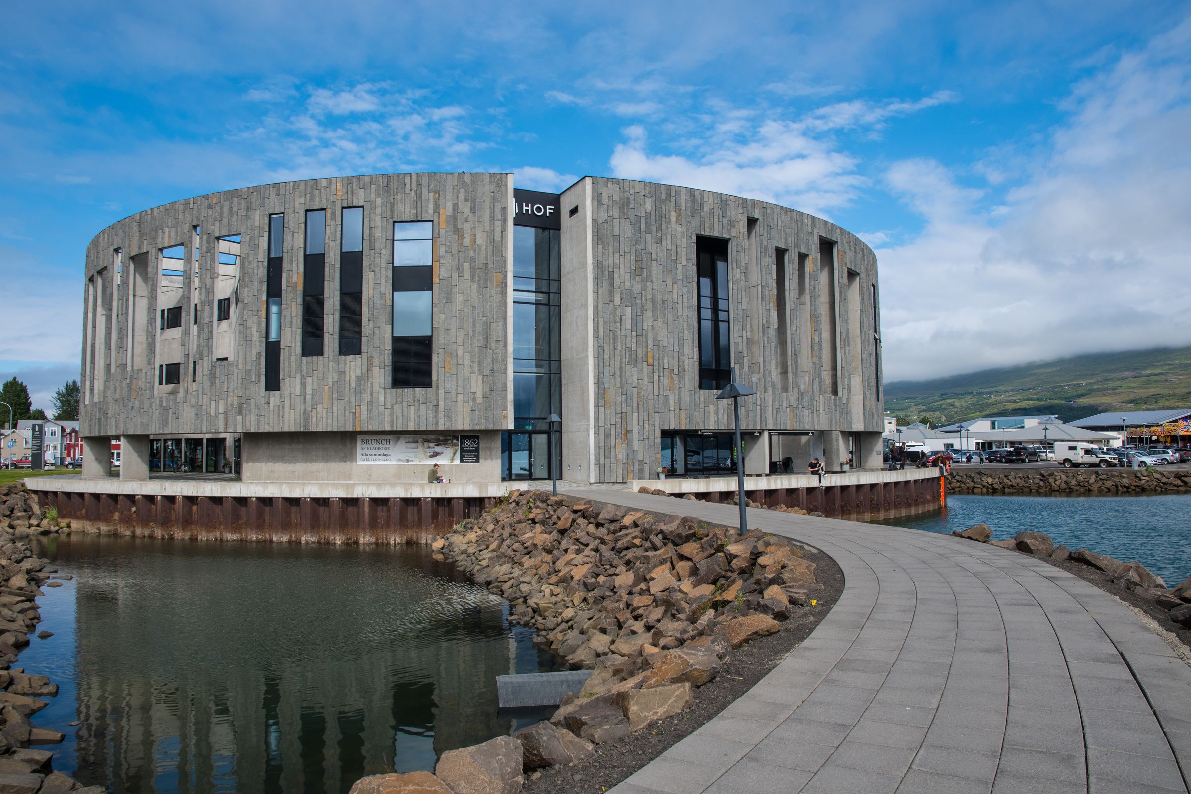 Un moderno edificio curvo de piedra gris con "HOF" en su fachada, ubicado junto a un puerto con un paseo pavimentado.