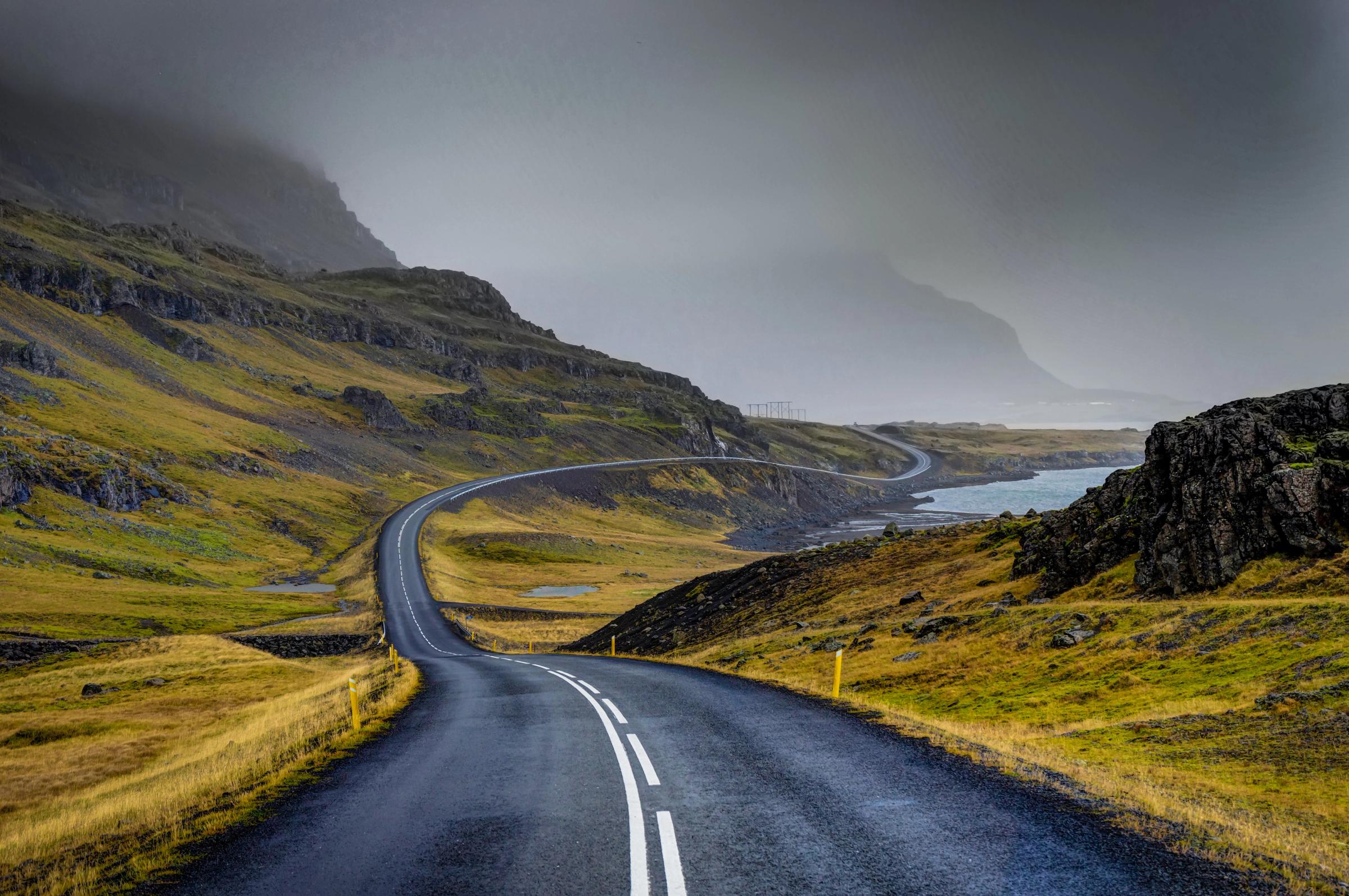 A winding coastal road hugs green hills and a bay under a cloudy sky.