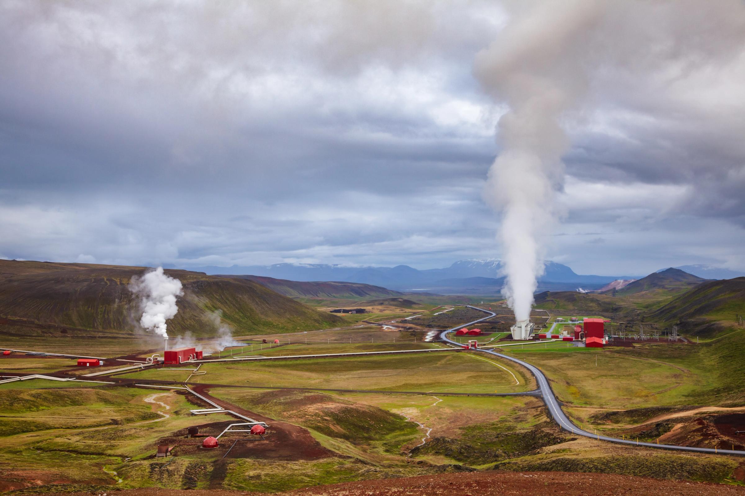 Geothermal power plant in a green valley, with two plumes of steam rising from its buildings under a cloudy sky.