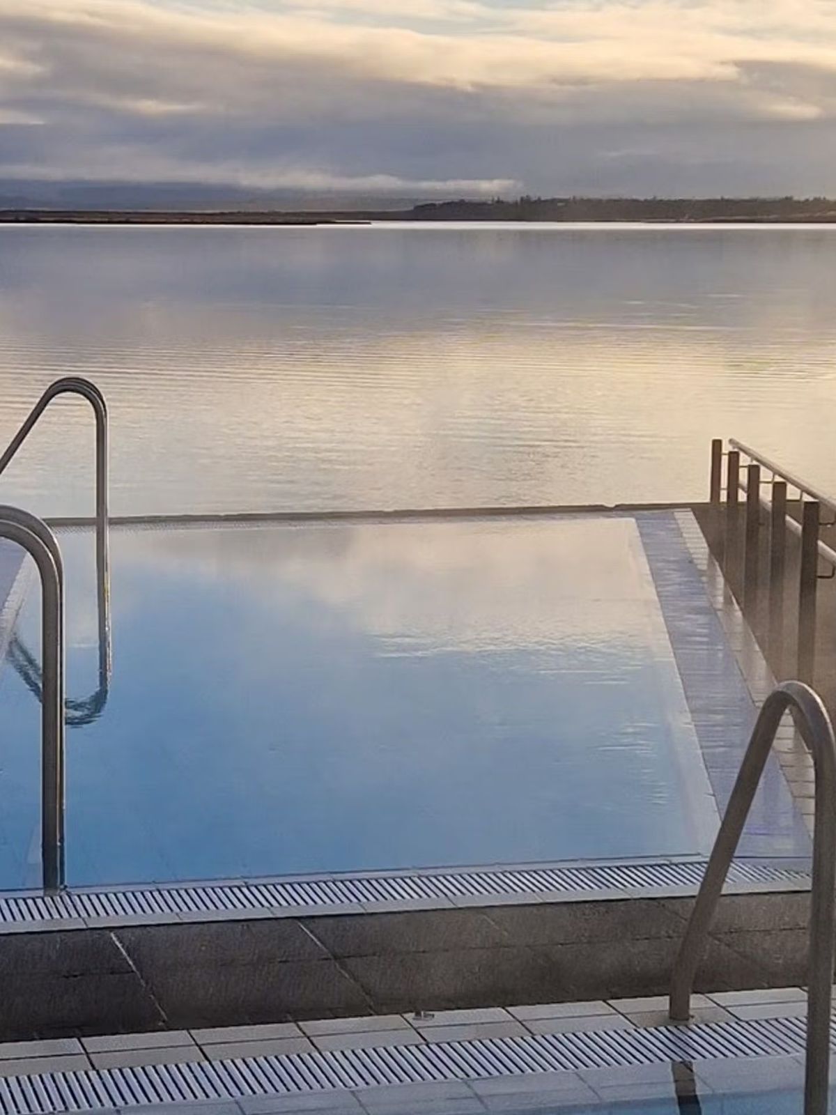 Piscina al aire libre con vapor con vista a un lago tranquilo bajo un cielo nublado.