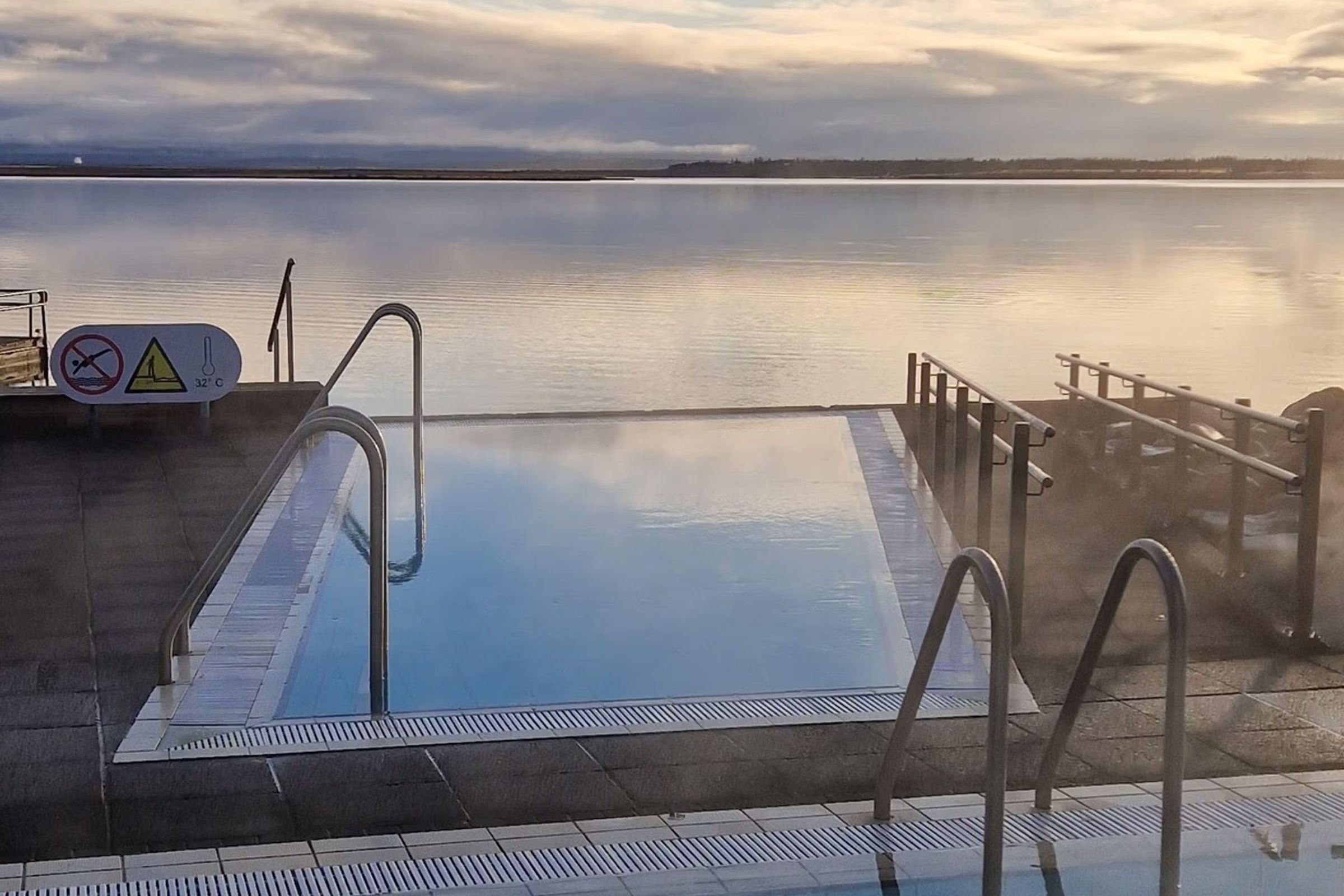 Una piscina infinita al aire libre, humeante, junto a un lago tranquilo bajo un cielo nublado.