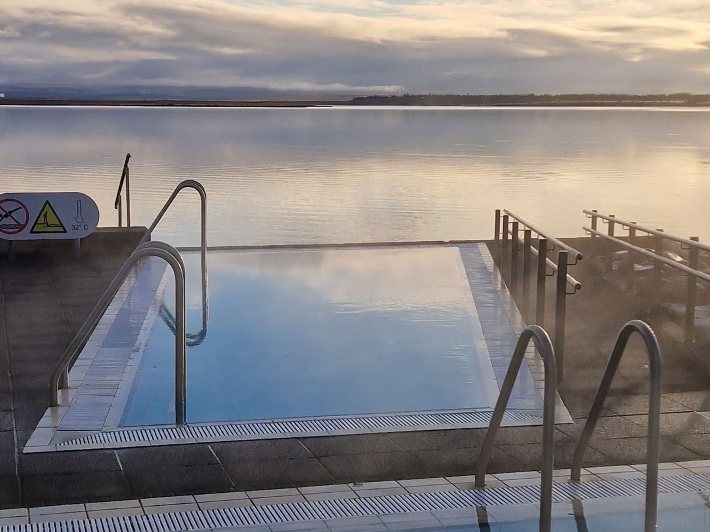 Steaming blue geothermal pool next to a calm body of water under a cloudy sky.