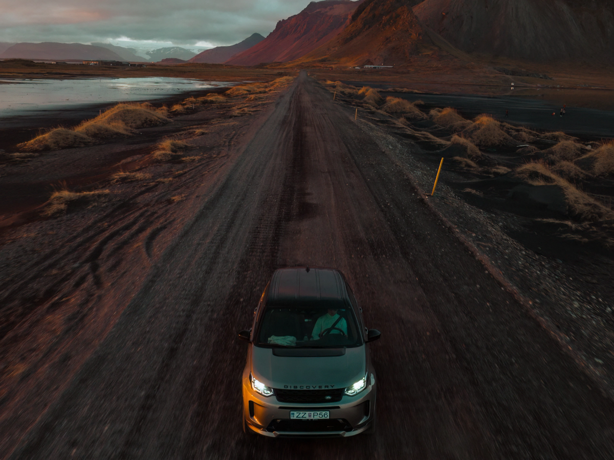 Discovery Sport aus der Vogelperspektive auf einer Schotterstraße Ein Land Rover Discovery Sport 4x4 fährt auf einer Schotterstraße, vor ihm liegen Berge.