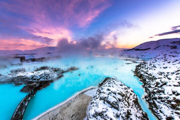 Steaming blue geothermal lagoon with bathers surrounded by snow, under a vibrant pink and purple sunset sky.