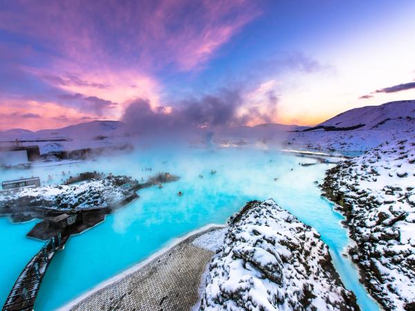 Geothermal blue lagoon with bathers and steam, surrounded by snow under a vibrant pink and purple sky.