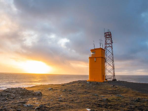 Orange lighthouse and communication tower on a rocky coast at sunset.