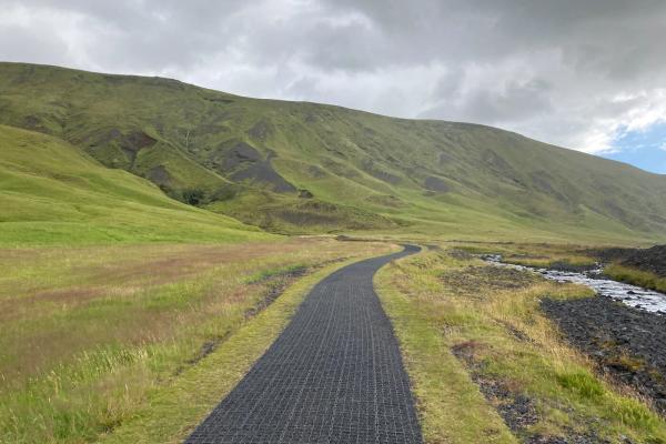 Winding dark path through a green valley with a stream, leading to rolling hills under a cloudy sky.