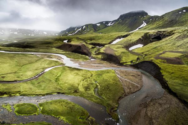 an aerial view of a river running through a grassy valley surrounded by mountains in the highlands in iceland.