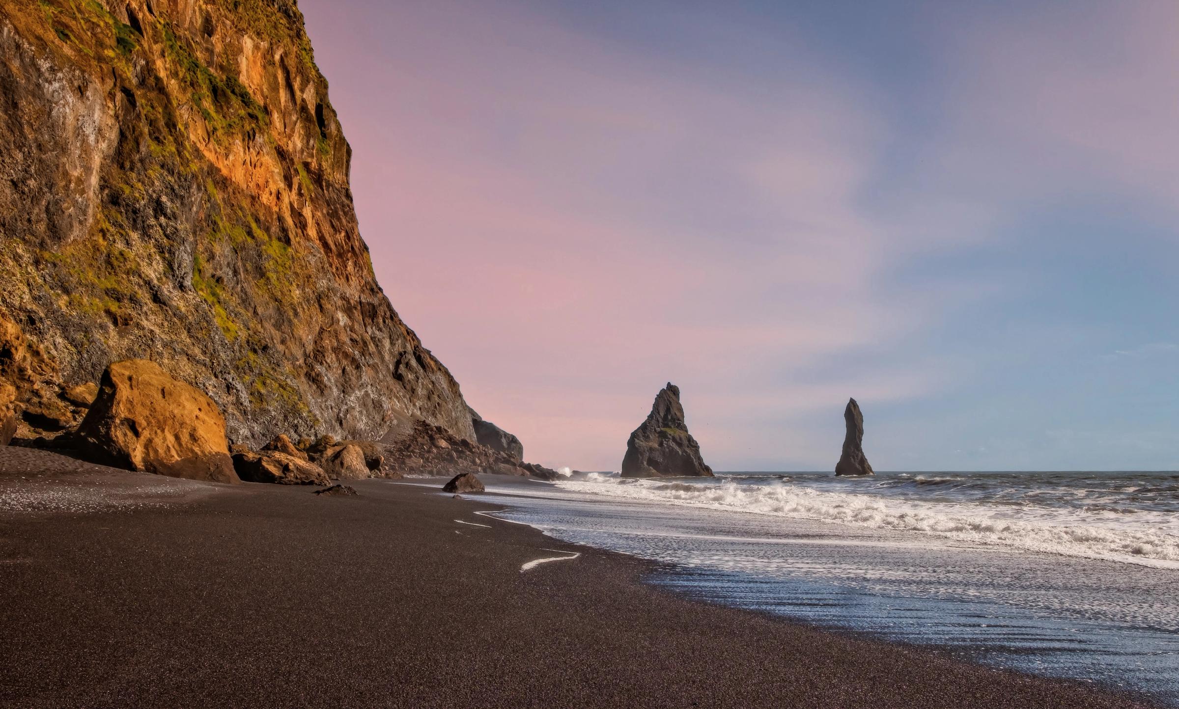 Black sand beach with a cliff, two sea stacks in the ocean, and a pink and blue sky.