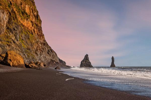 Black sand beach with a cliff, two sea stacks in the ocean, and a pink and blue sky.