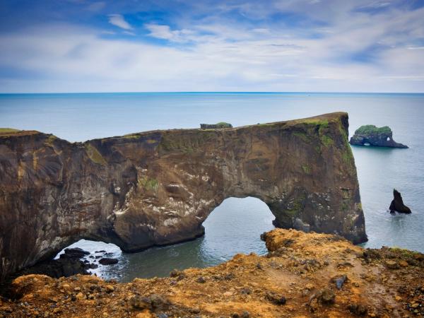 A natural rock arch extends into the sea under a cloudy sky, with smaller rock formations in the distance.
